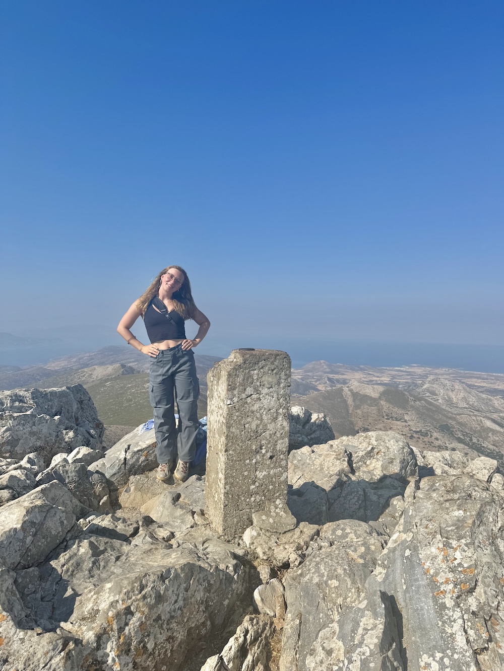 Ivy McDonald atop a mountain in Greece, standing on boulders with a bright blue sky in the background.